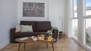 Un salón moderno con un sofá marrón, una mesa de centro con alimentos para el desayuno y un ventanal con vistas a edificios.