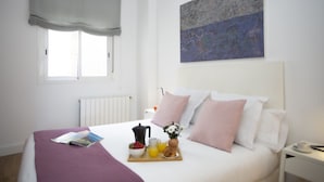 Una cama bien hecha con una bandeja de desayuno, una ventana con cortina blanca y una obra de arte abstracta en la pared.