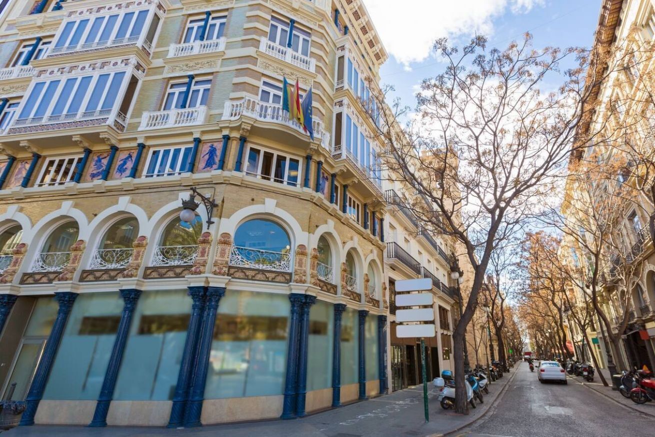 Um edifício histórico com varandas elaboradas e grandes janelas, situado numa rua arborizada.