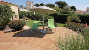 An outdoor patio with two green lounge chairs and a large umbrella, surrounded by well-maintained gardens and a building with a tiled roof in the background. - Calypso - Javea, Costa Blanca (Javea)