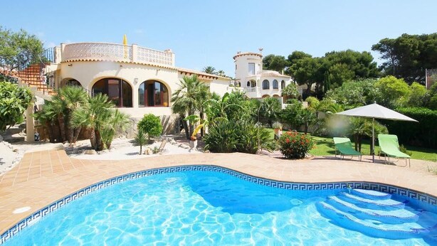 A swimming pool with a tiled edge, surrounded by a landscaped garden with palm trees and a building with arched windows and a tower. - Calypso - Javea, Costa Blanca (Javea)