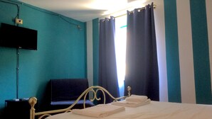 Ein Zimmer mit Türkiswänden, einem Bett mit weißer Bettwäsche, einem schwarzen Stuhl, einem an der Wand befestigten Flachbildfernseher und einem Fenster mit dunklen Vorhängen.