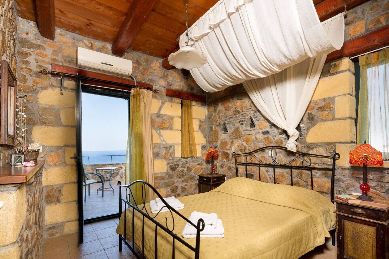 A room with a bed, a stone wall, a balcony, and a view of the sea.