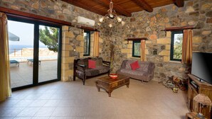 A stone-walled room with a wooden ceiling, a television, a sofa, a coffee table, and a view of the sea through a glass door.