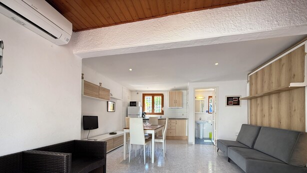Una sala moderna con un sofá gris, un banco de mimbre oscuro y una zona de comedor con mesa y sillas. Se ve una cocina al fondo con armarios de madera y un ventanal.