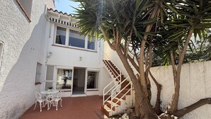 Un edificio de dos pisos con una escalera que lleva a un balcón. Hay un patio con una mesa y sillas, y un árbol en primer plano.