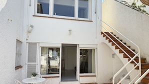 Un edificio de dos pisos con un patio que incluye una mesa y sillas blancas, una escalera y una puerta que da a un espacio interior.