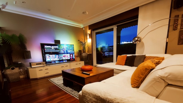 Una sala de estar moderna con un sofá blanco, una mesa de centro de madera y un televisor de pantalla plana mostrando un servicio de streaming.