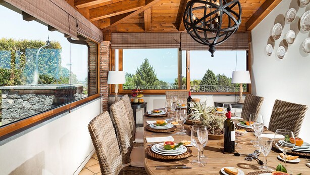 Un comedor con mesa de madera dispuesta para una comida, sillas de mimbre y vistas a zonas verdes.