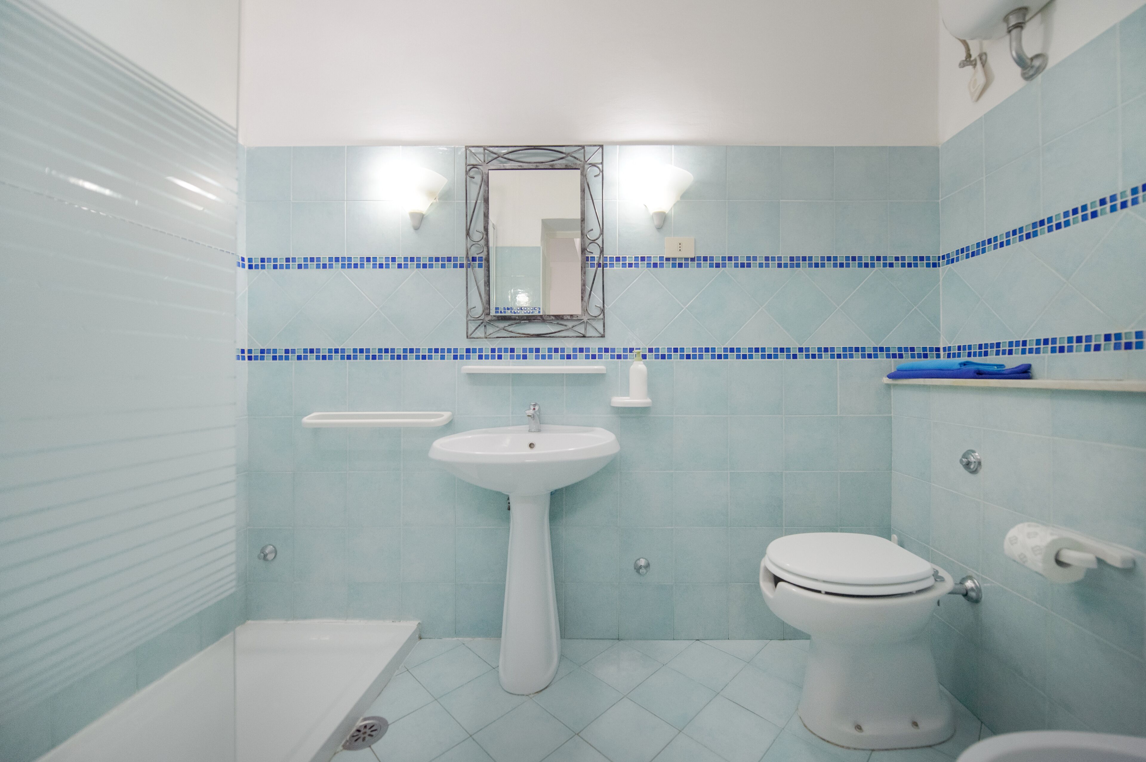 Une salle de bain avec un lavabo, des toilettes et une baignoire, tous de couleur blanche. Un miroir est placé au-dessus du lavabo, et une étagère contient des articles de toilette.