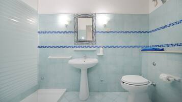 Une salle de bain avec un lavabo, des toilettes et une baignoire, tous de couleur blanche. Un miroir est placé au-dessus du lavabo, et une étagère contient des articles de toilette.