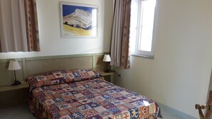 Een bed met een beddekleed met patroon, twee bedlampjes, een schilderij aan de muur en een raam met gordijnen.
