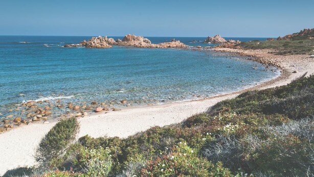 Een kustlandschap met helderblauw water, een zandstrand en rotsformaties.