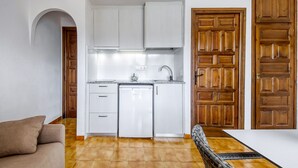 A compact kitchen with white cabinets, a dishwasher, and a sink. There is a wooden door with a geometric pattern, a brown sofa, and a dining table. - JARDINS I 22 Roses (Rosas / Roses)