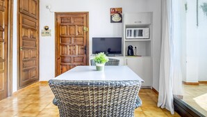 A room with a white table, wicker chair, wooden door, TV, and a shelf with a coffee machine. - JARDINS I 22 Roses (Rosas / Roses)