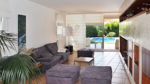 Ein modernes Wohnzimmer mit einem grauen Sofa, zwei Hocker, einem Couchtisch und einem großen Fenster mit Blick auf einen Pool.