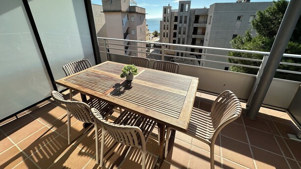 Ein Balkon mit einem Holztisch und Stühlen, einem kleinen Blumentopf und Blick auf Gebäude sowie das Meer.