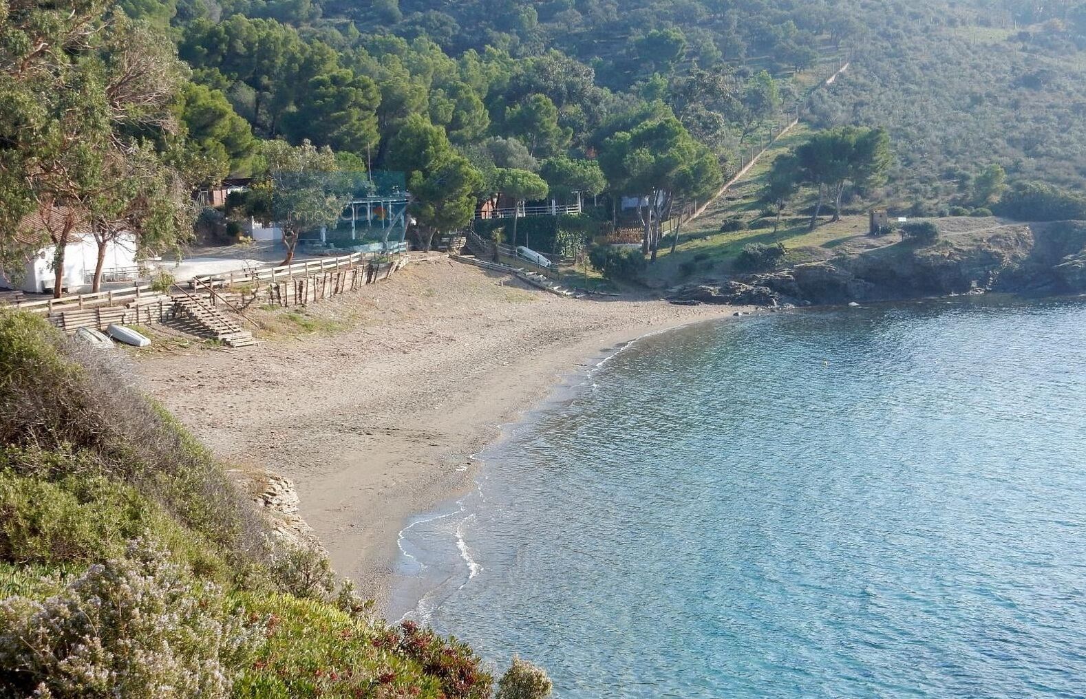 Un paisaje costero con una playa arenosa, una cerca de madera y un fondo de árboles y colinas.