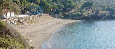 Un paisaje costero con una playa arenosa, una cerca de madera y un fondo de árboles y colinas.