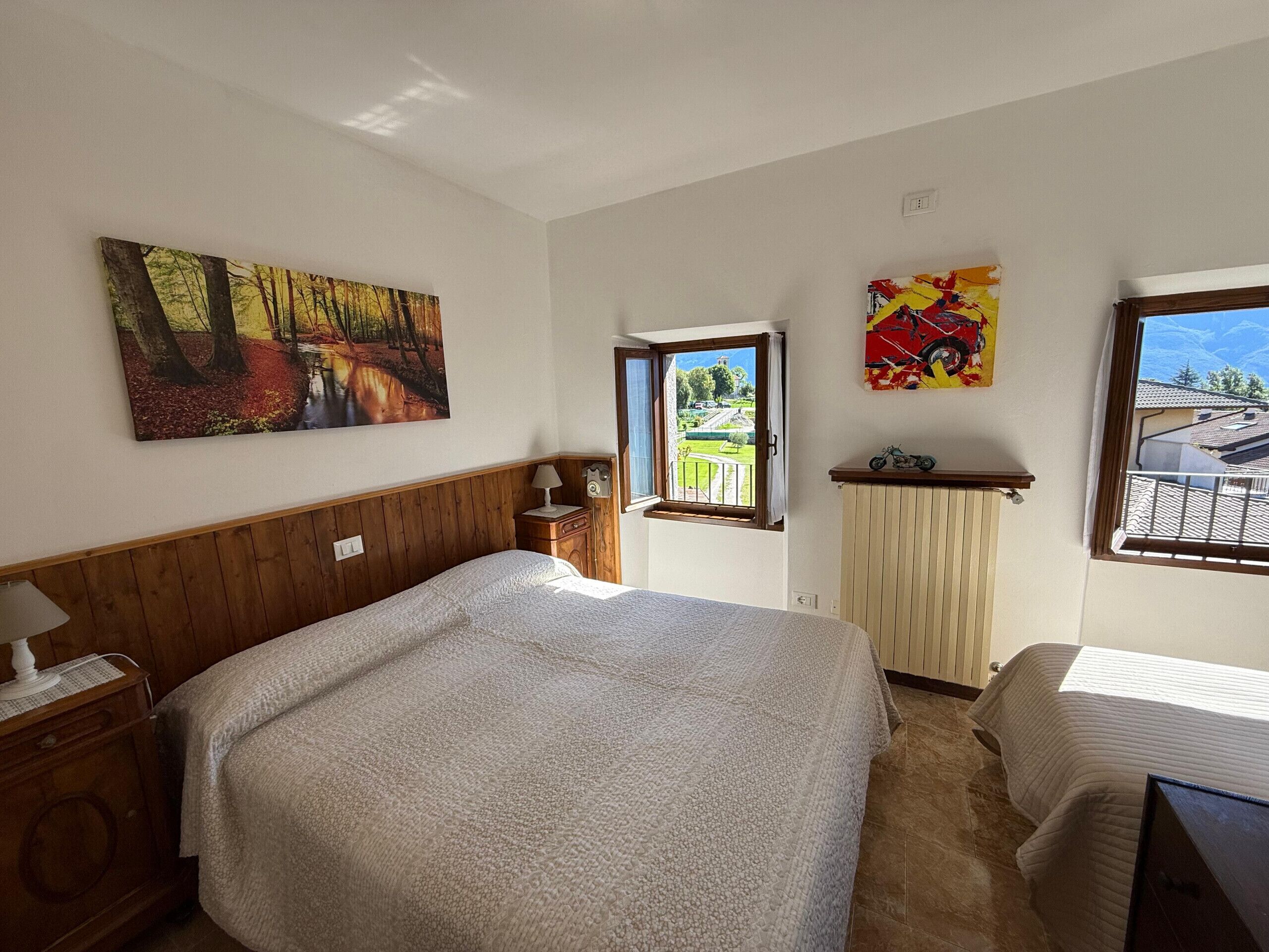 A bedroom with a bed, a nightstand, a window with a view of a garden, and a painting on the wall.
