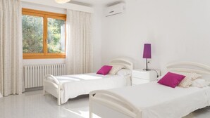 A bright bedroom with two beds, a window with curtains, a radiator, an air conditioner, and a bedside lamp.