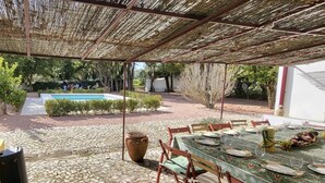 An outdoor dining area with a table set for four, surrounded by stone flooring and a pergola overhead.