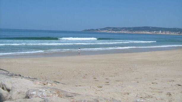 A beach with clear skies, calm waves, and a distant cityscape.