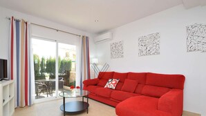 Una sala de estar con un sofá rojo, una mesa de centro de vidrio y un televisor sobre un mueble. Hay una puerta corrediza de vidrio que da a un área exterior con una mesa y sillas.