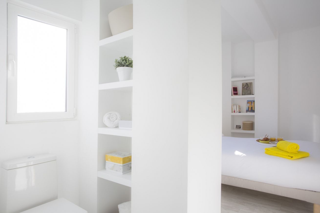 A modern, white-themed bathroom with a toilet, shelves with toiletries, and a window.