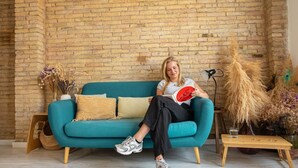 Une personne est assise sur un canapé de couleur turquoise, avec un mur de briques derrière elle. Une petite table en bois se trouve à côté du canapé, et un vase contenant des plantes séchées est posé sur le côté.