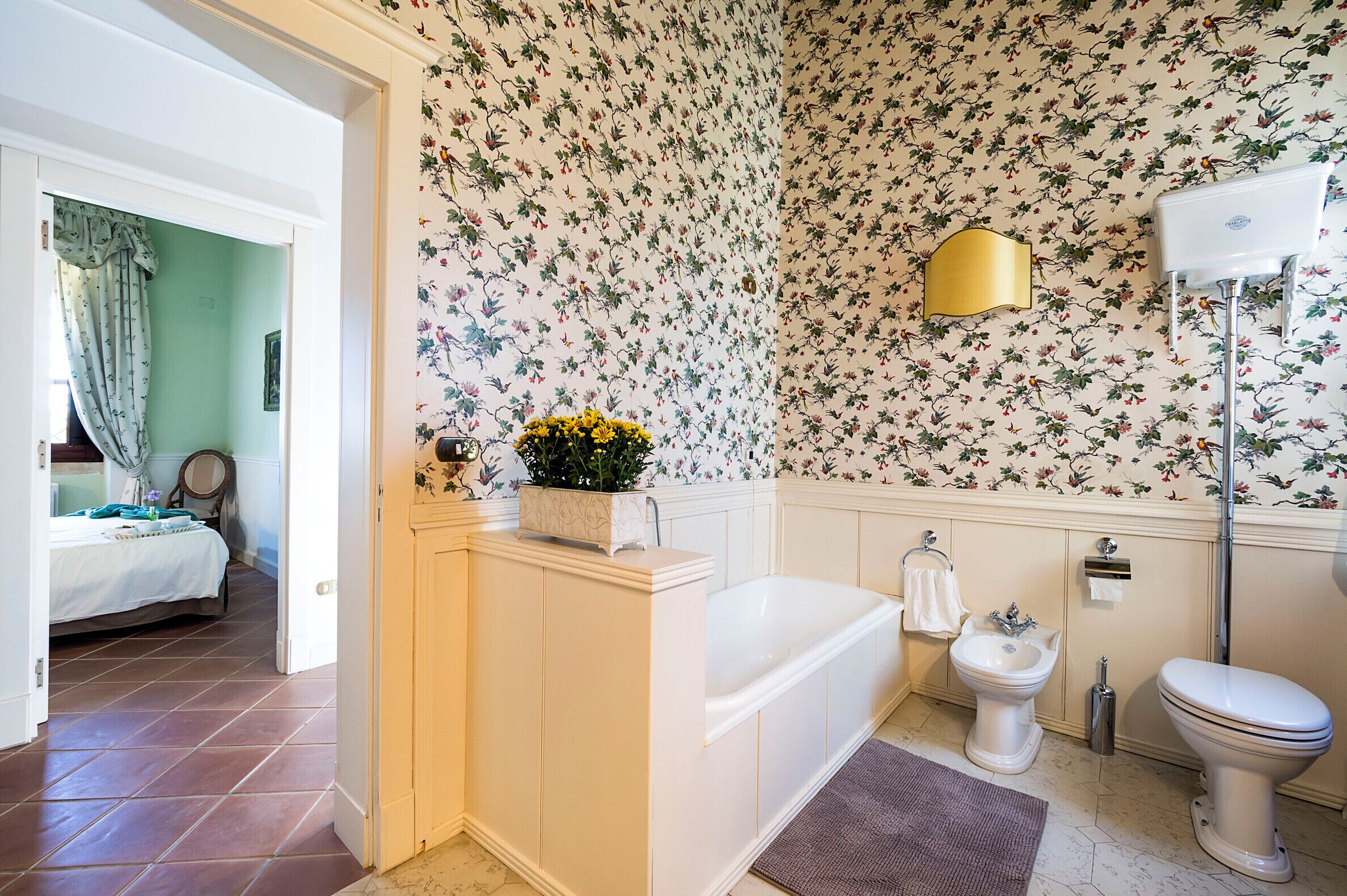 Ein Badezimmer mit Badewanne, Toilette und Bidet. Die Wände sind mit floralem Papier tapeziert und eine goldene Wandleuchte ist angebracht. Ein Bett ist im angrenzenden Raum zu sehen.