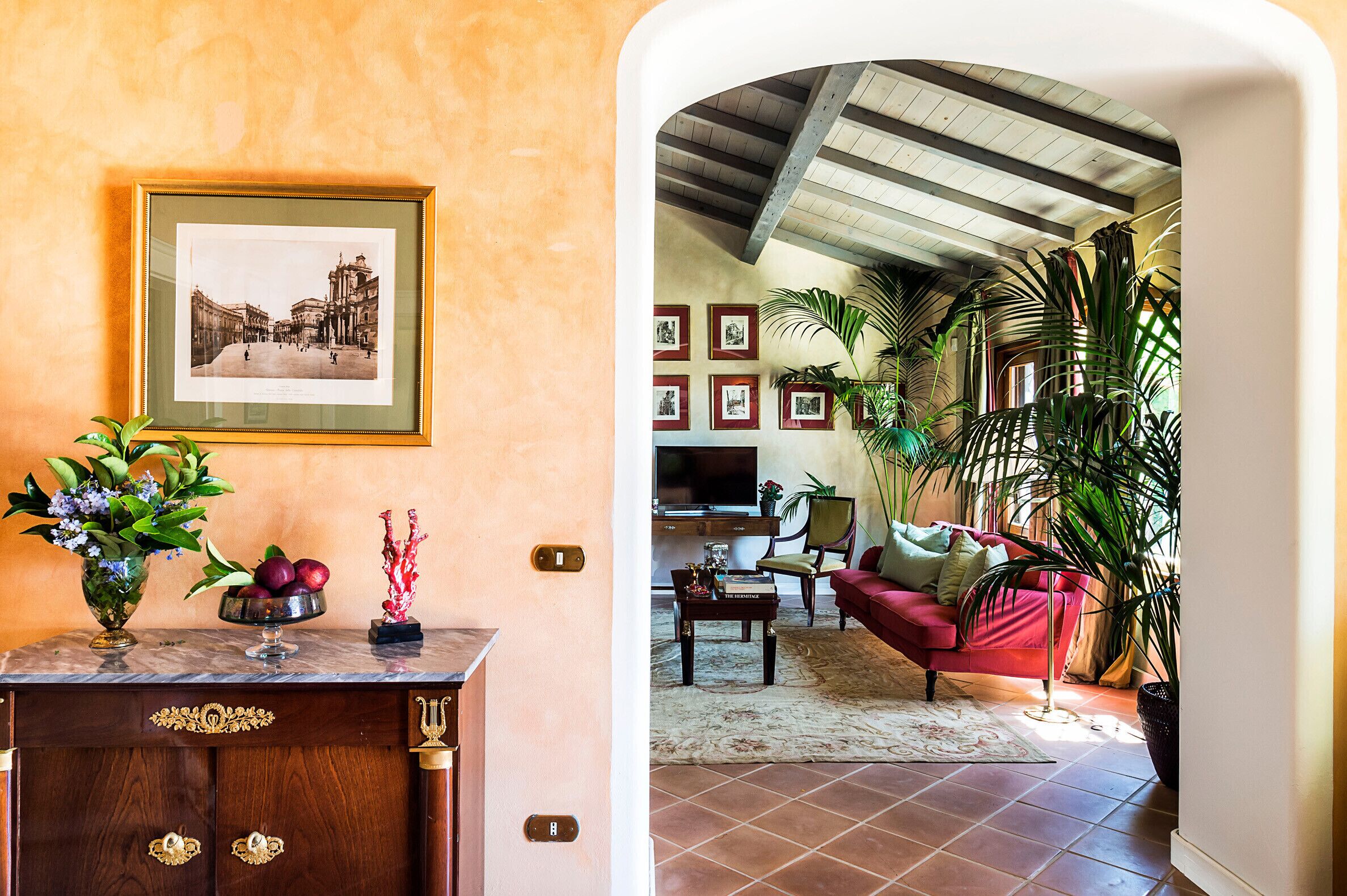 Un salon avec un canapé rouge, une table basse et un téléviseur. Des tableaux encadrés ornent le mur, et un vase rempli de fleurs est posé sur la commode.
