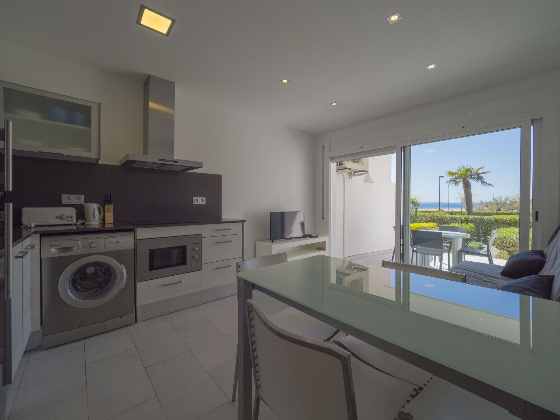 A modern kitchen with a washing machine, oven, and microwave. There is a dining area with a table and chairs, and a view of the sea through large windows.