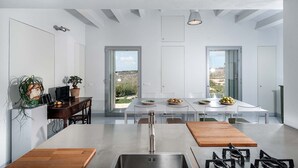 A modern kitchen with a central island, white cabinetry, and large windows offering a view of the outdoors. - Designer country villa with pool in Ragusa, Sicily (Ragusa)