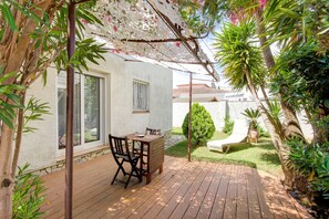 A wooden deck with a table and chairs, surrounded by lush greenery and a white building with large windows. - VILLA LA SELVA 26 Roses (Rosas / Roses)