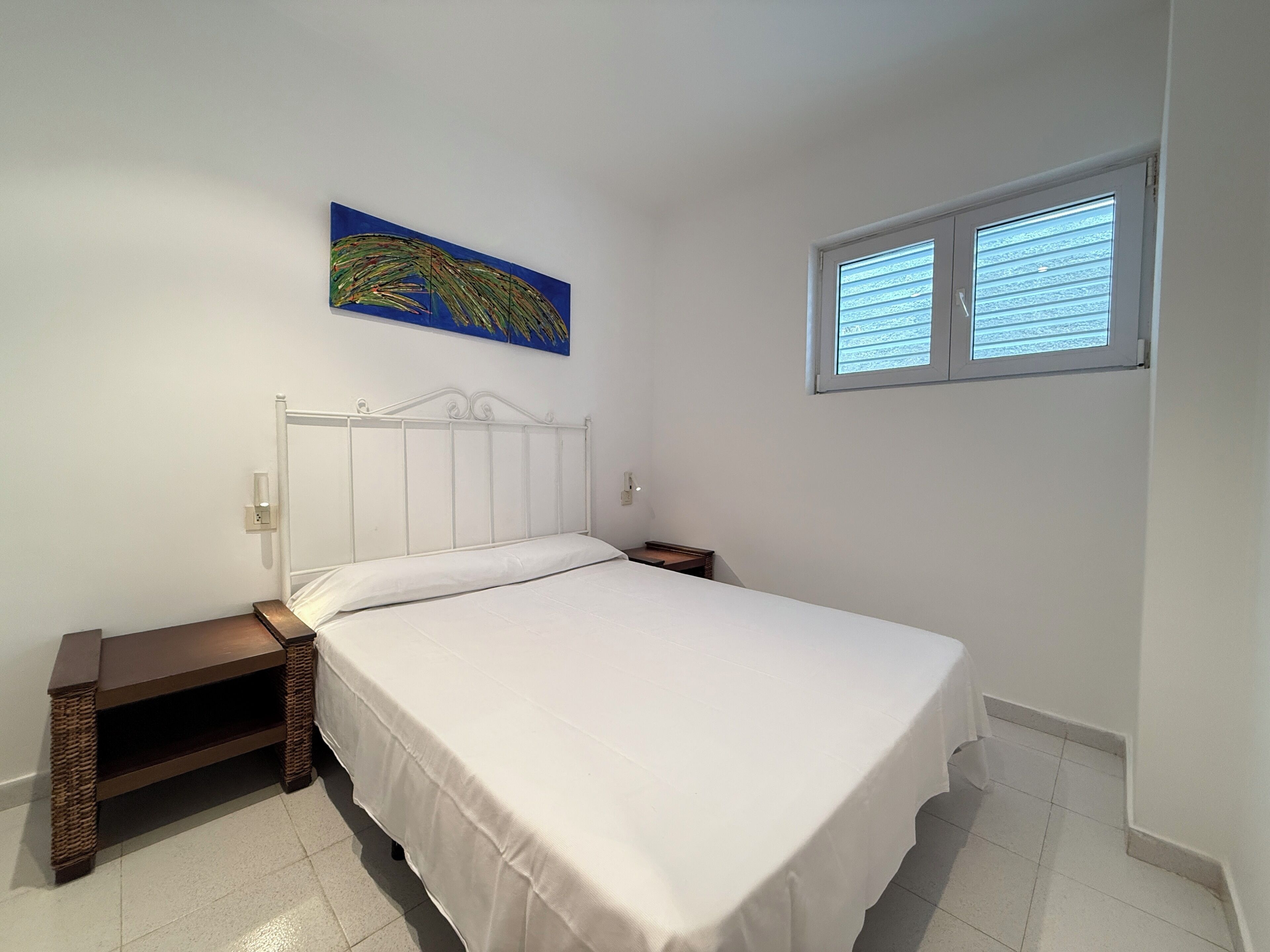 A bedroom with a bed, a nightstand, a small table, a painting, and a window with blinds.