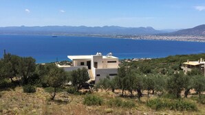 A modern building with a flat roof and solar panels, surrounded by trees and overlooking a coastal area with mountains in the distance. - Luxury villa with private heated pool and stunning sea views. (Kalamata)