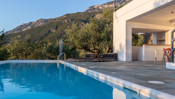 A swimming pool with a mountain backdrop, a modern outdoor seating area, and a stone wall. - Luxury villa with private heated pool and stunning sea views. (Kalamata)