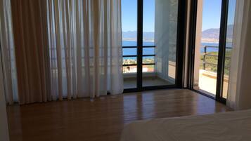 A room with a large sliding glass door leading to a balcony with a view of the sea and mountains.
