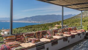 A covered outdoor seating area with colorful woven cushions, overlooking a coastal city and mountains. - Luxury villa with private heated pool and stunning sea views. (Kalamata)
