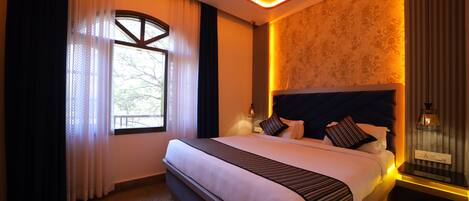 A hotel room with a large bed, a window with curtains, a nightstand with a lamp, and a wall with patterned wallpaper.