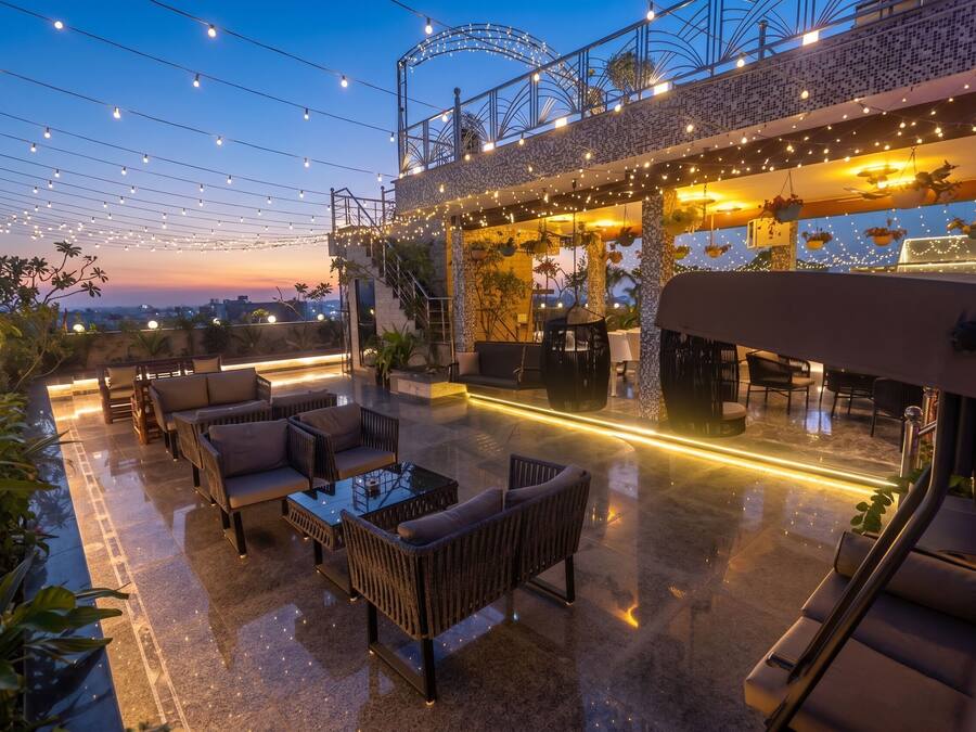 Un espacio al aire libre con mobiliario de mimbre y plantas colgantes, bajo una zona cubierta con luces de cadena.