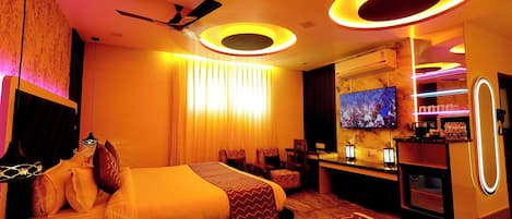 A hotel room with a bed, seating area, TV, minibar, and a ceiling fan.