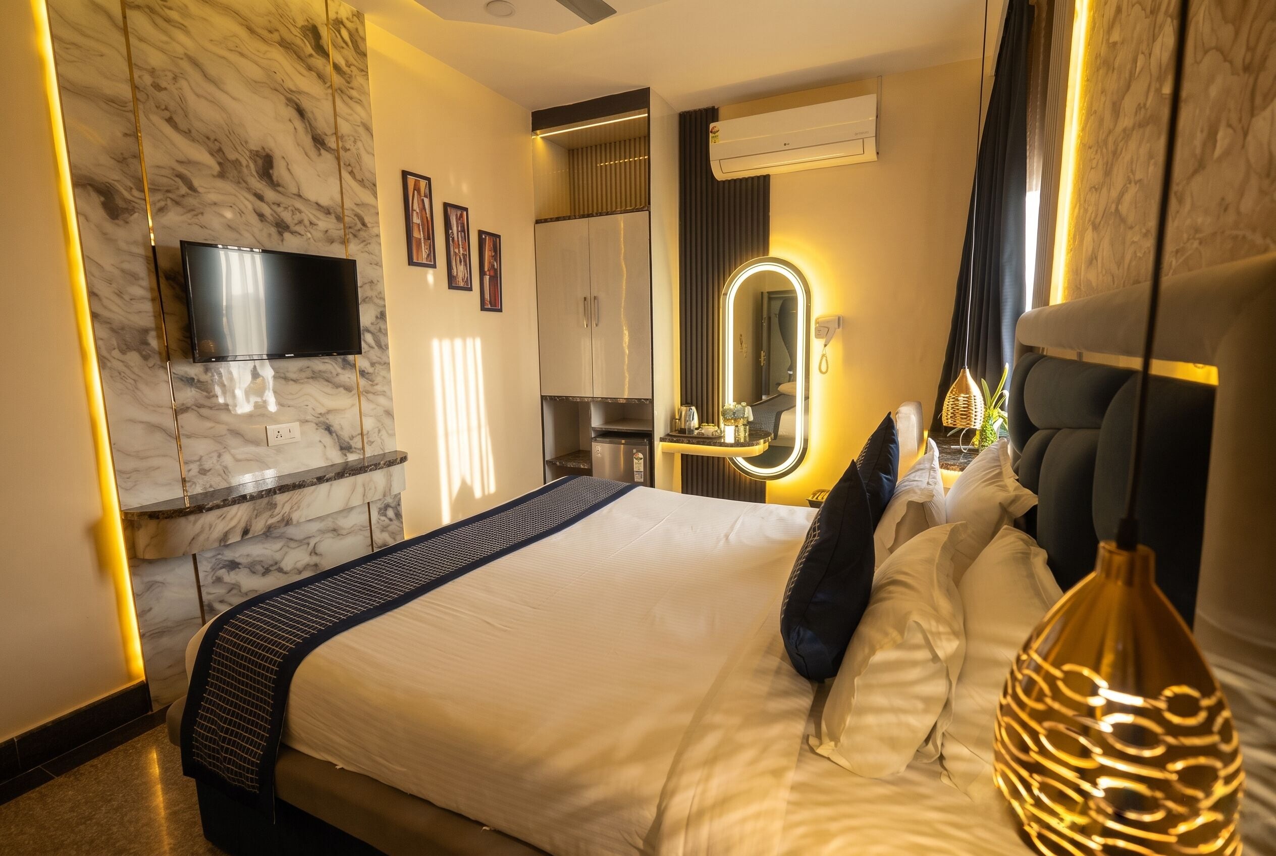 A modern hotel room with a marble wall, a flat-screen TV, a large bed with a headboard, a bedside table, and a mirror with a light.