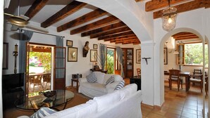 A cozy living room with a sofa, a glass table, and a ceiling fan. There is a dining area with chairs and a table, and a kitchen visible in the background. - Can Teulada, private pool Pollensa 12 persons (Pollensa)