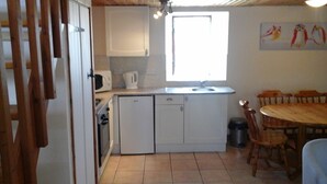 A compact kitchen with white cabinets, a microwave, and a sink. There is a dining table with chairs and a window above the sink. - Rose Gite Le Cosquer Saint Connec (Saint-Connec)
