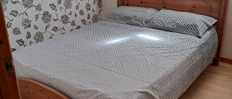 A wooden bed with a patterned bedspread and pillows, a wooden headboard, and a floral wallpaper pattern on the wall.