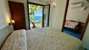Un dormitorio con cama, una puerta de madera, una mesita con silla y vista al océano desde una puerta abierta.