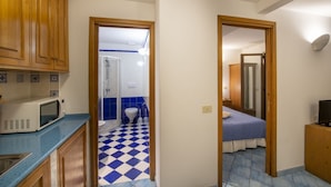 Cocina compacta con armarios de madera y un microondas, baño con azulejos azules y un dormitorio con cama y televisor.