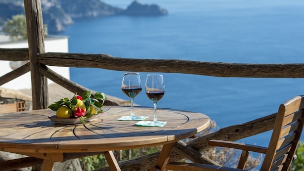 Una mesa de madera con dos copas de vino y un frutero en un balcón con vistas al mar.